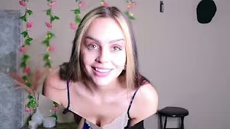 Young woman with straight blonde hair wearing a white silk camisole, standing in front of a wall decorated with pink floral garlands and string lights