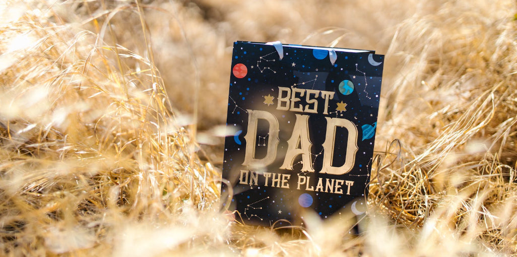 A 'Best Dad on the Planet' greeting card with space-themed design featuring planets and constellations, nestled in golden dry grass