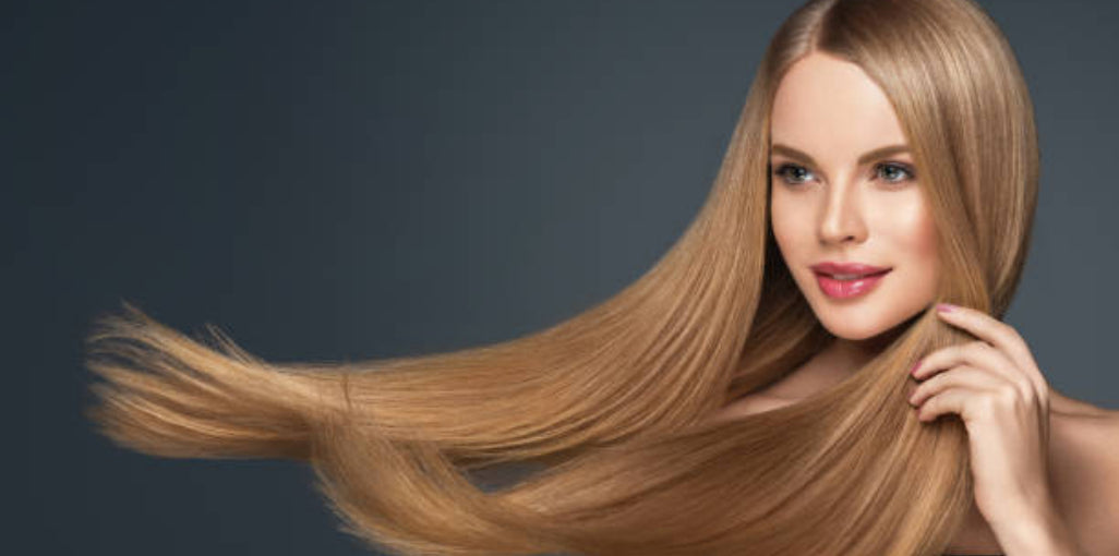 A smiling woman with long, smooth, flowing blonde hair, gently touching her hair as it billows