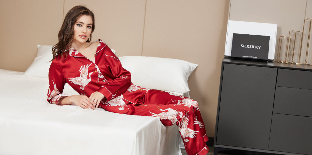 Woman wearing luxurious red silk pajamas with white crane pattern, sitting on a bed with SILKSILKY brand packaging visible on a side table