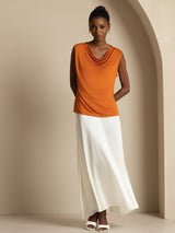 [Orange] Orange cowl-neck top with a white maxi skirt in a standing front view, hands behind the body and white sandals visible. 005