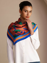 [Clay Elephant] Coral pink square scarf with blue, green and brown animal motifs, blue border, styled front, at neck, hand near chin. 004