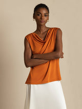 [Orange] Front upper-body view of an orange cowl-neck sleeveless top with crossed arms and a white maxi skirt. 003