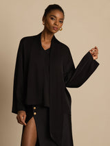 [Black] Black draped-neck blouse with a black skirt in a front crop to thighs, one arm crossing the torso and the other lowered. 004