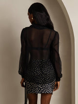 [Black] Rear studio view of a model wearing a sheer black long-sleeve tie-neck blouse over a black-and-white patterned skirt. 002