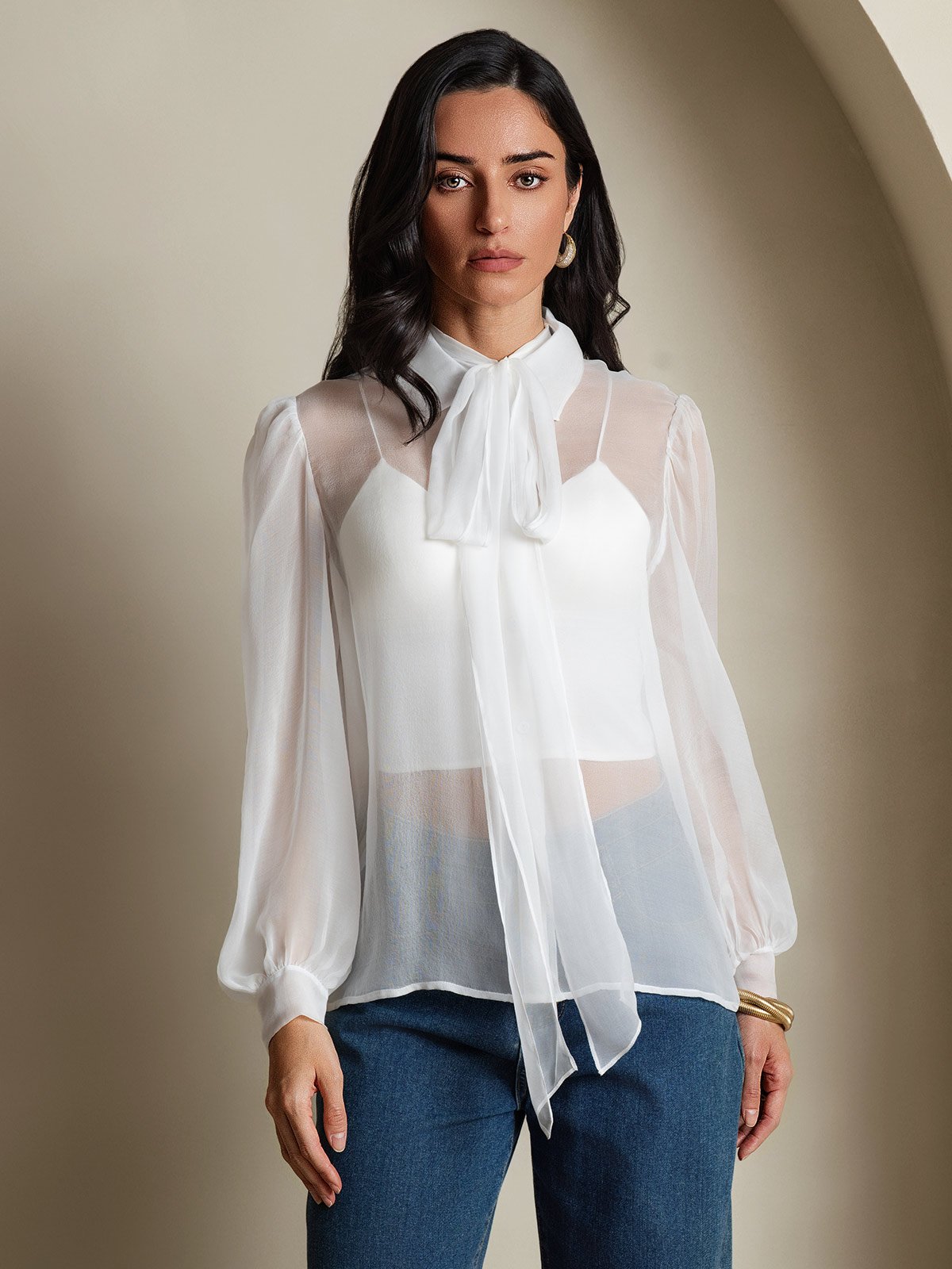 [White] Studio view of a model wearing a sheer white long-sleeve tie-neck blouse with blue trousers. 001