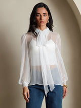 [White] Studio view of a model wearing a sheer white long-sleeve tie-neck blouse with blue trousers. 001