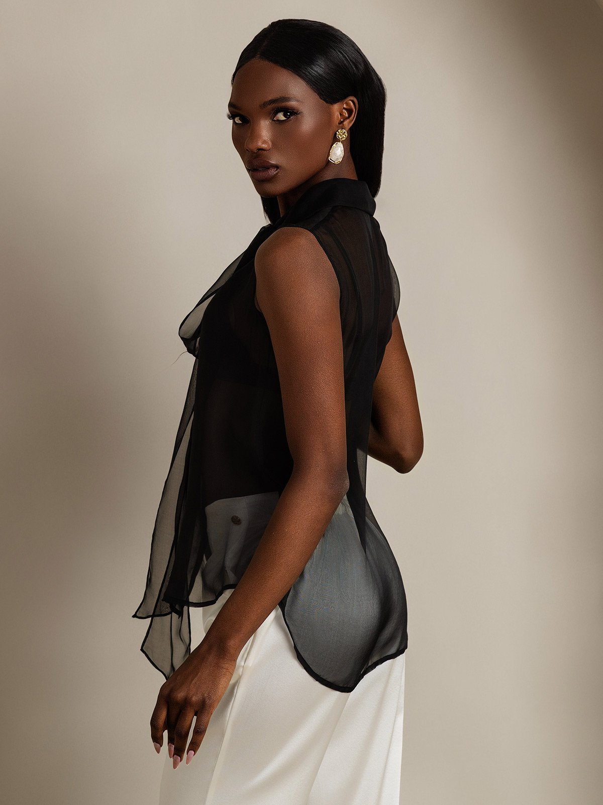 [Black] Rear studio view of a model wearing a sheer black sleeveless bow-neck blouse with white trousers. 002