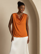 [Orange] Orange cowl-neck top with a white maxi skirt is shown from the front at mid length as one arm lifts beside the body. 001