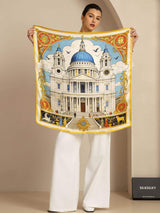 [P107] Studio view of a model holding a square scarf with an ornate multicolor architectural print and gold border. 002