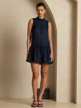 [Dark Blue] Navy drop-waist mini dress with ruffle hem in a standing front view, both arms resting down and sandals visible. 003