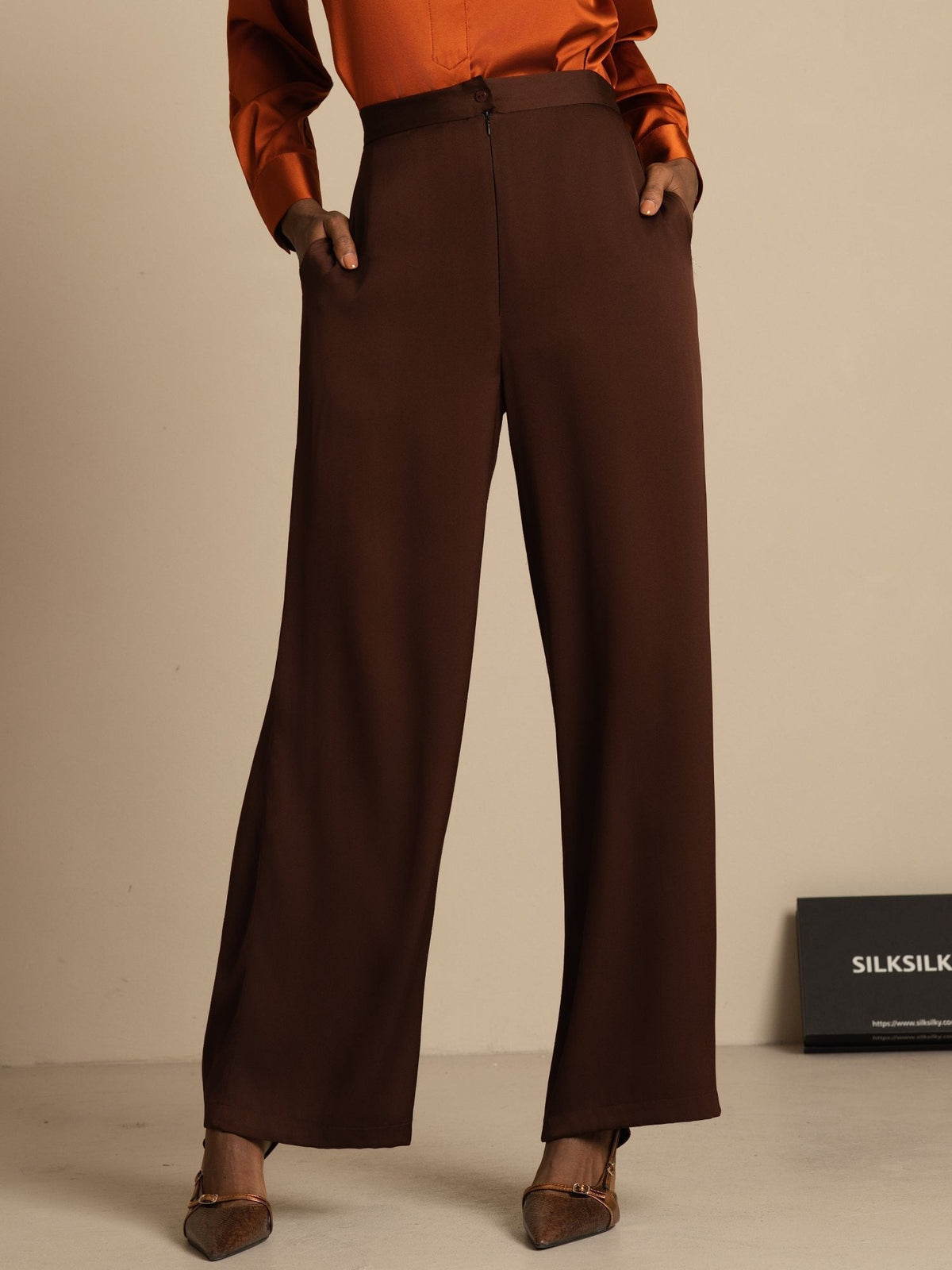 [Bitter Chocolate] Front three-quarter view: brown high-rise wide-leg trousers with a rust-orange blouse, with one arm bent. 001