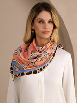 [Pink Cat] Coral pink animal-print square scarf with blue-and-black patterned border, styled front, over shoulders, hand at edge. 004