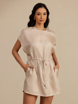 [Tan] light tan cap-sleeve short dress with drawstring waist and pockets, waist-up view, model with one hand in a pocket. 001
