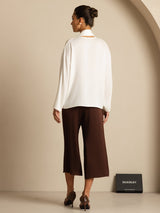 [White] Rear studio view of a model wearing a white draped-neck long-sleeve blouse with dark brown wide-leg trousers. 002